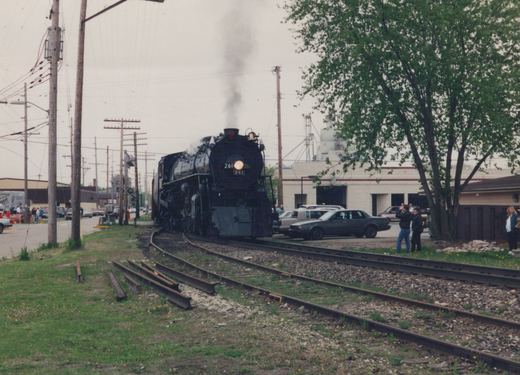A black steam locomotive numbered 261 emits smoke as it travels on tracks beside a street with parked 1990s cars and people watching.