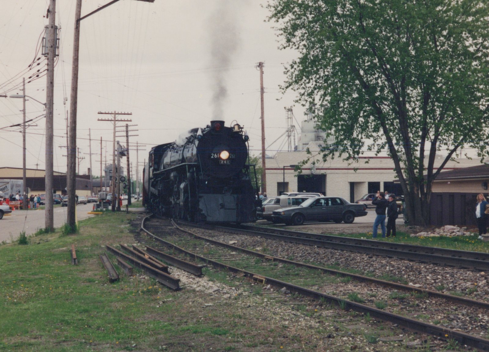 A black steam locomotive numbered 261 emits smoke as it travels on tracks beside a street with parked 1990s cars and people watching.