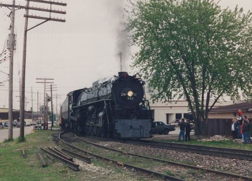 A black steam locomotive numbered 261 emits smoke as it travels along tracks beside a grassy area, with spectators nearby and utility poles in the background.