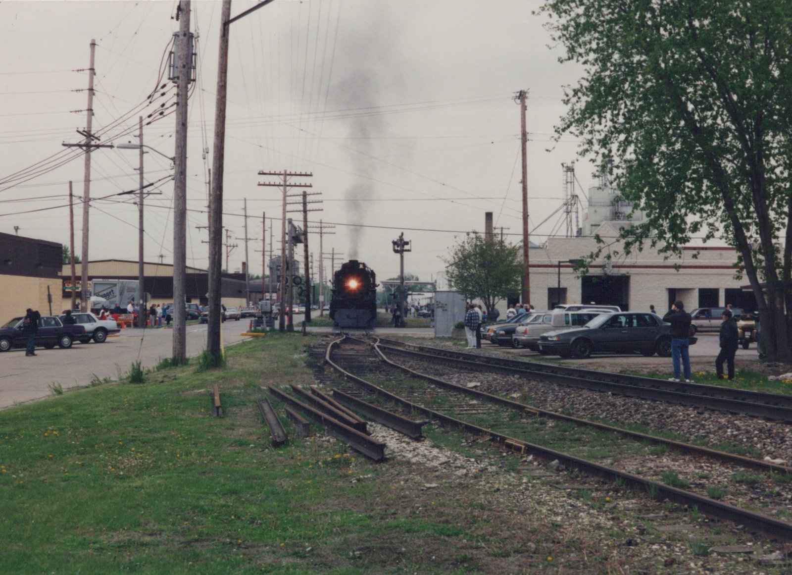 A black steam locomotive with its headlight on approaches a grade crossing on a cloudy day, surrounded by spectators and parked cars along the roadside.