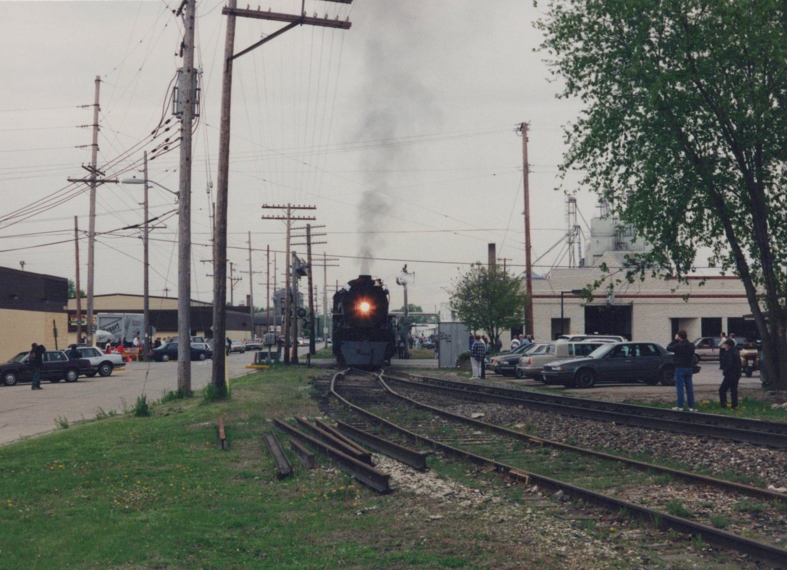 A black steam locomotive with the number 261 on its front is moving toward the camera on railroad tracks, with spectators and parked cars nearby under an overcast sky.