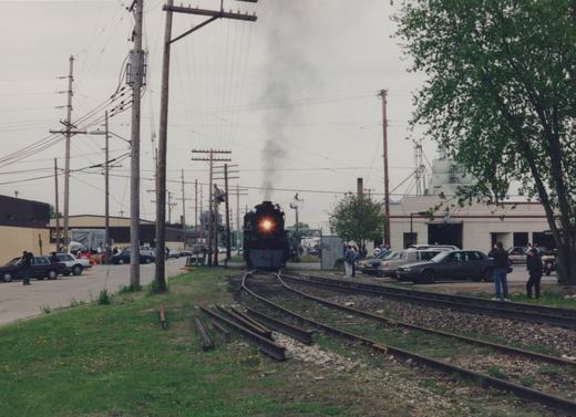 A black steam locomotive with the number 261 on its front is moving toward the camera on railroad tracks, with spectators and parked cars nearby under an overcast sky.