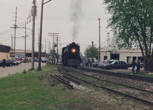 A black steam locomotive numbered 261 releases smoke as it moves along tracks beside a street lined with cars and onlookers, with buildings and utility poles in the background.
