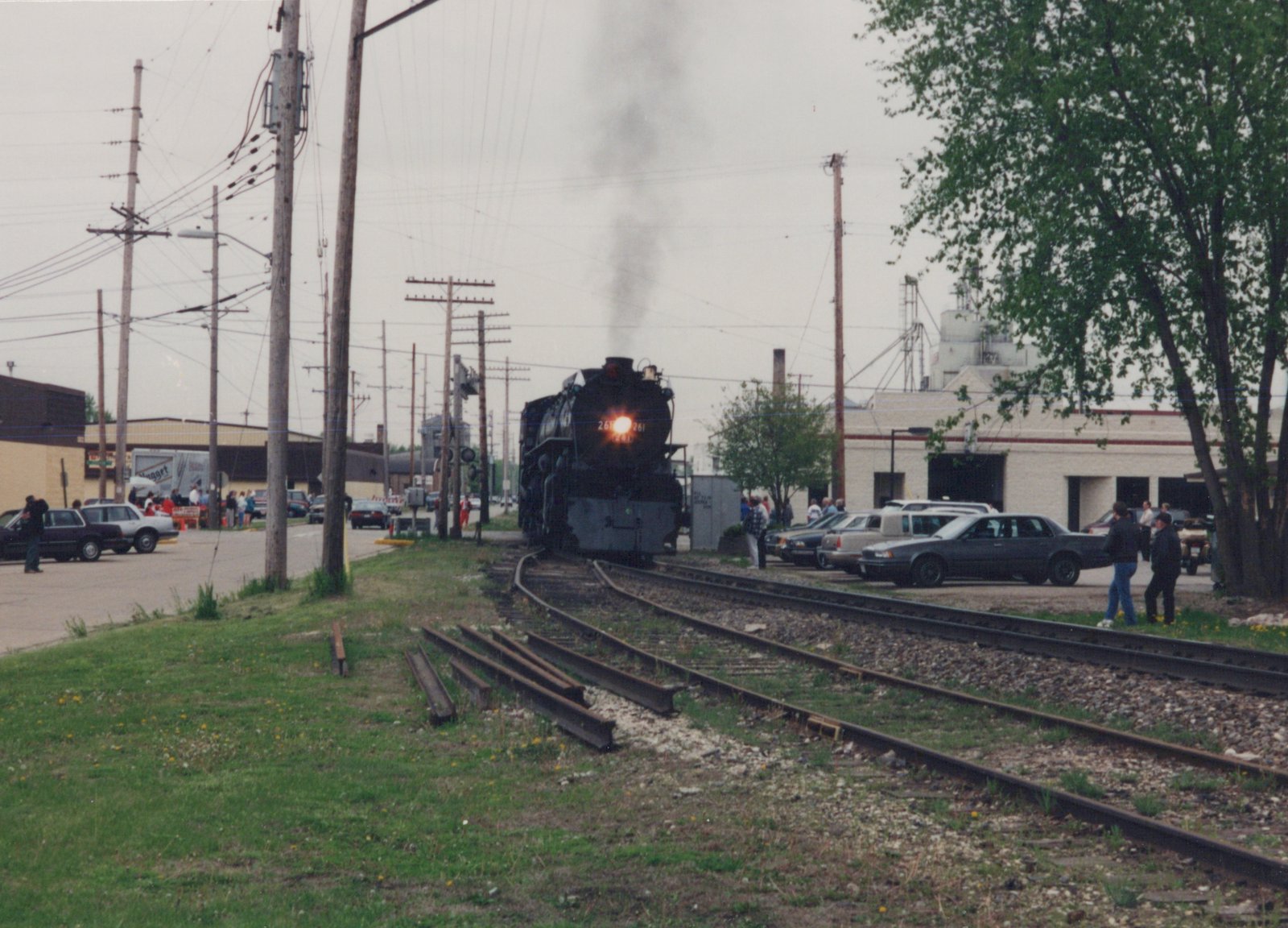 A black steam locomotive numbered 261 releases smoke as it moves along tracks beside a street lined with cars and onlookers, with buildings and utility poles in the background.