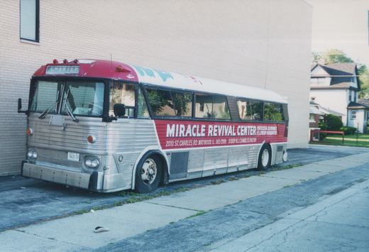 A silver and red charter bus for the Miracle Revival Center is parked on pavement next to a beige brick building and residential houses in daylight.