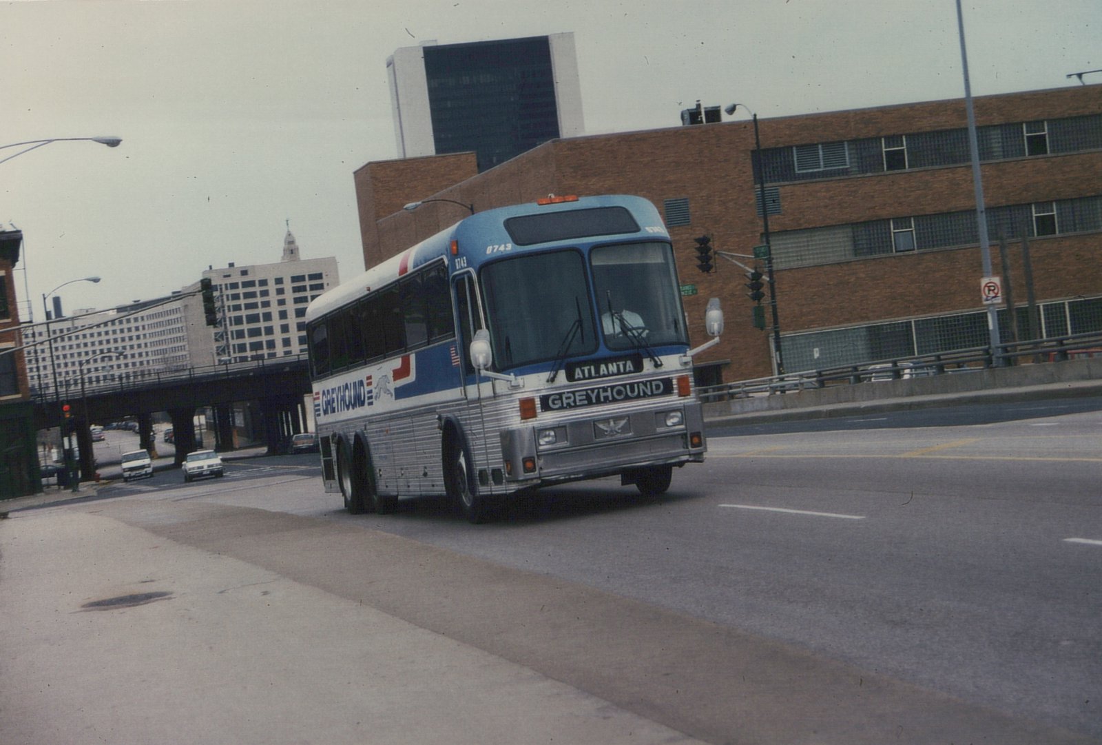 A large Greyhound bus with a blue and white livery, marked 'Atlanta', drives down a city street with buildings and other cars in the background.