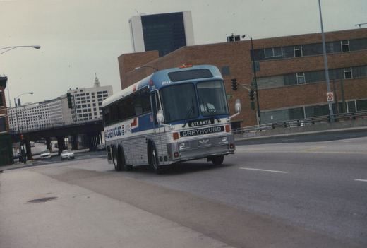 A large Greyhound bus with a blue and white livery, marked 'Atlanta', drives down a city street with buildings and other cars in the background.