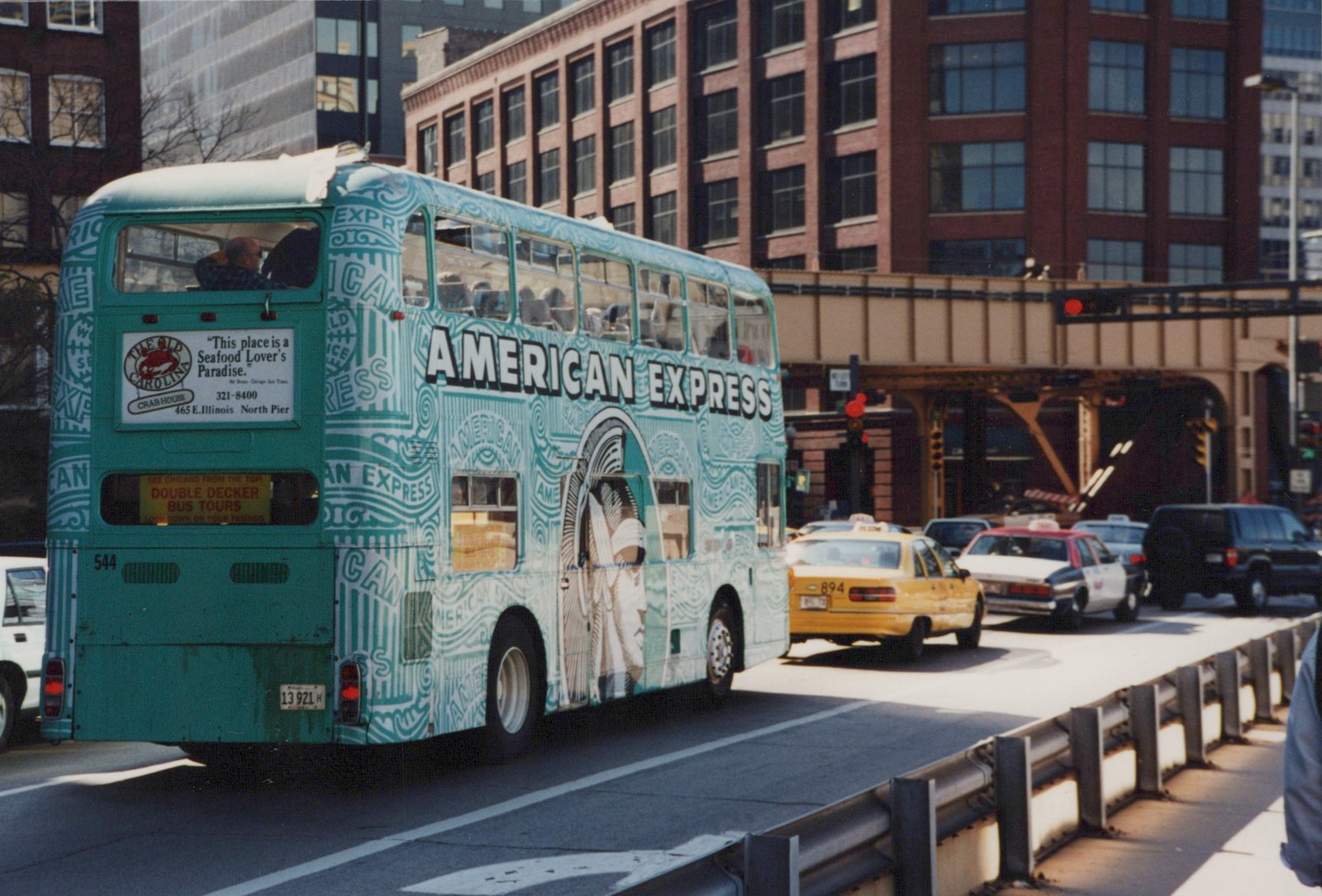 A teal and white double-decker tour bus with 'AMERICAN EXPRESS' lettering, number 544, is on a city street with a yellow taxi and other cars nearby, under an elevated structure.