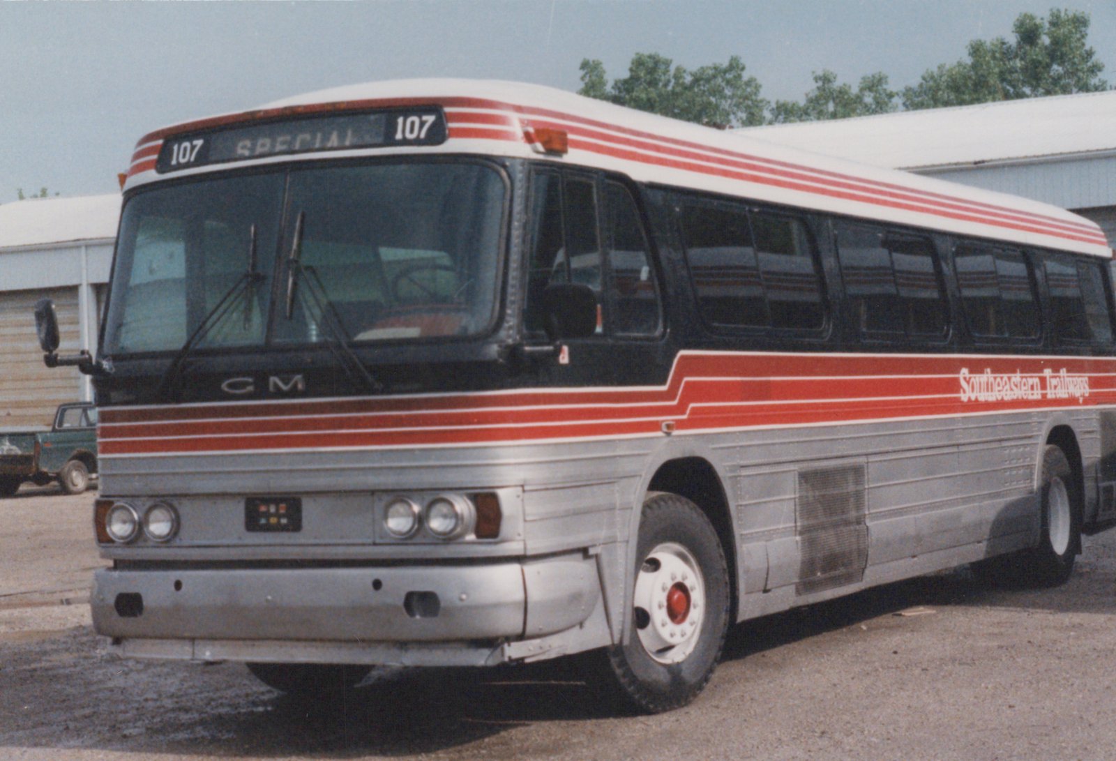A silver and red Southeastern Trailways motor bus, number 107, is parked on a gravel lot with a green pickup truck in the background.