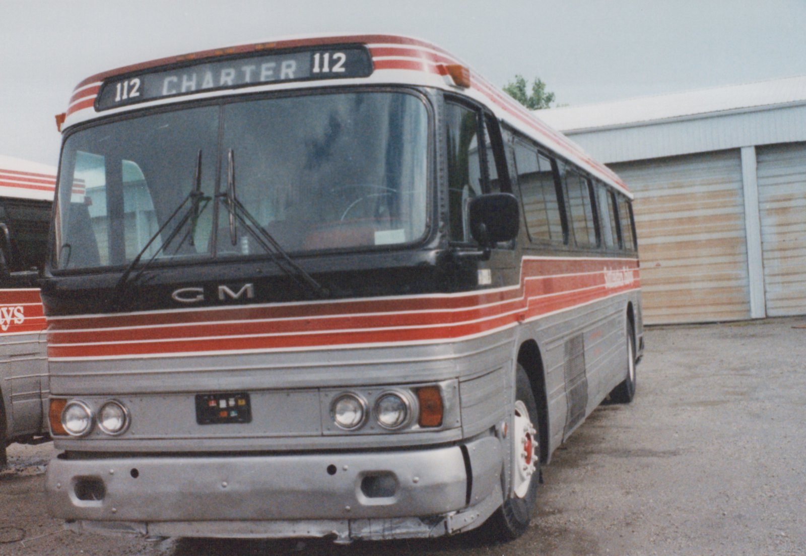 A silver and red striped GM New Look motor bus with fleet number 112, labeled 'CHARTER' on the front, parked in front of a garage with another bus partially visible to the left.