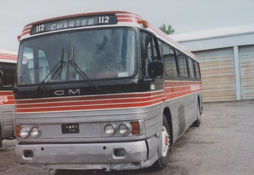 A silver and red striped GM New Look motor bus with fleet number 112, labeled 'CHARTER' on the front, parked in front of a garage with another bus partially visible to the left.