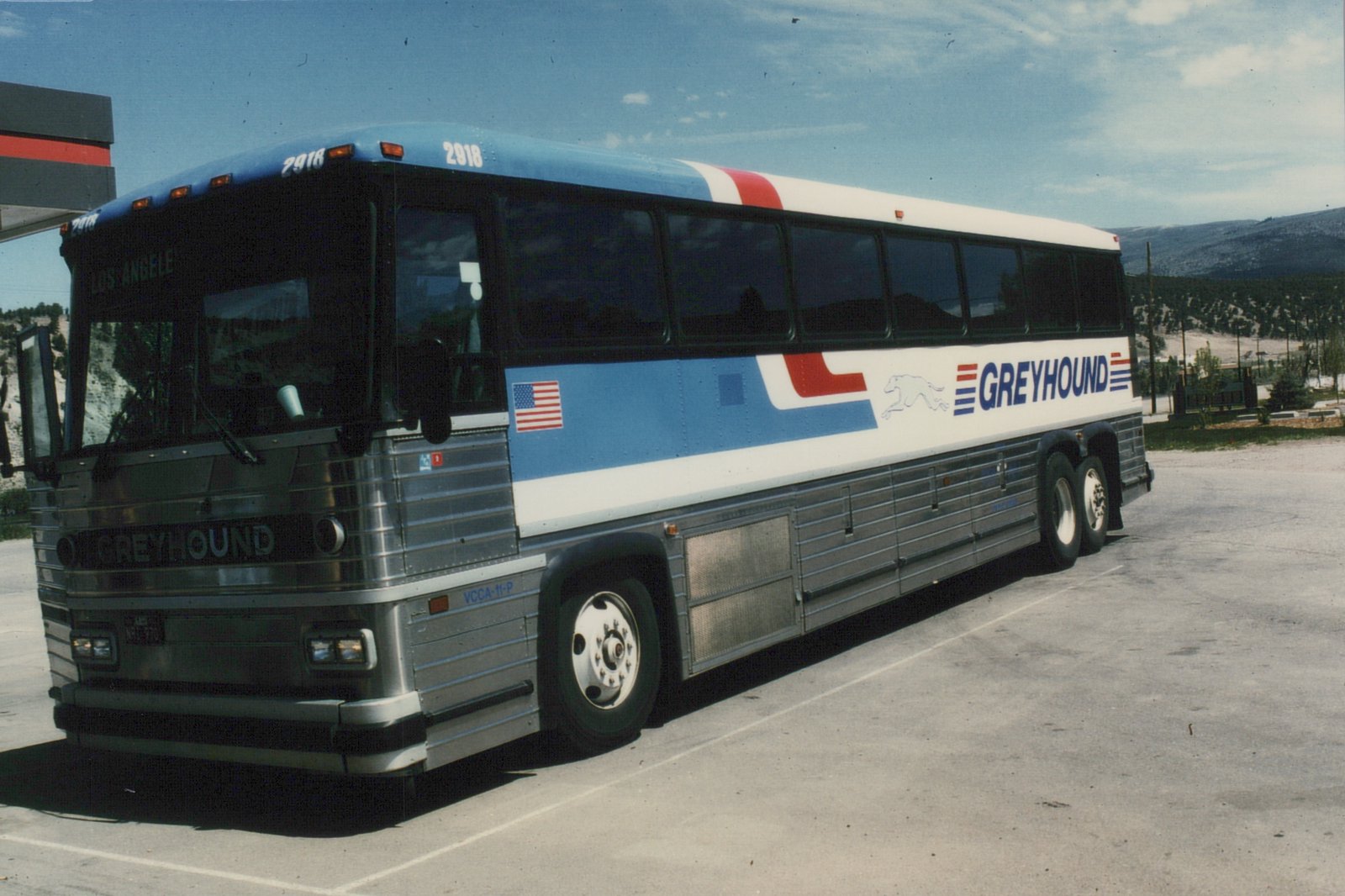 A side-front view of a Greyhound bus, number 2918, in its classic blue, white, and red livery, parked on a paved lot with mountains in the background.
