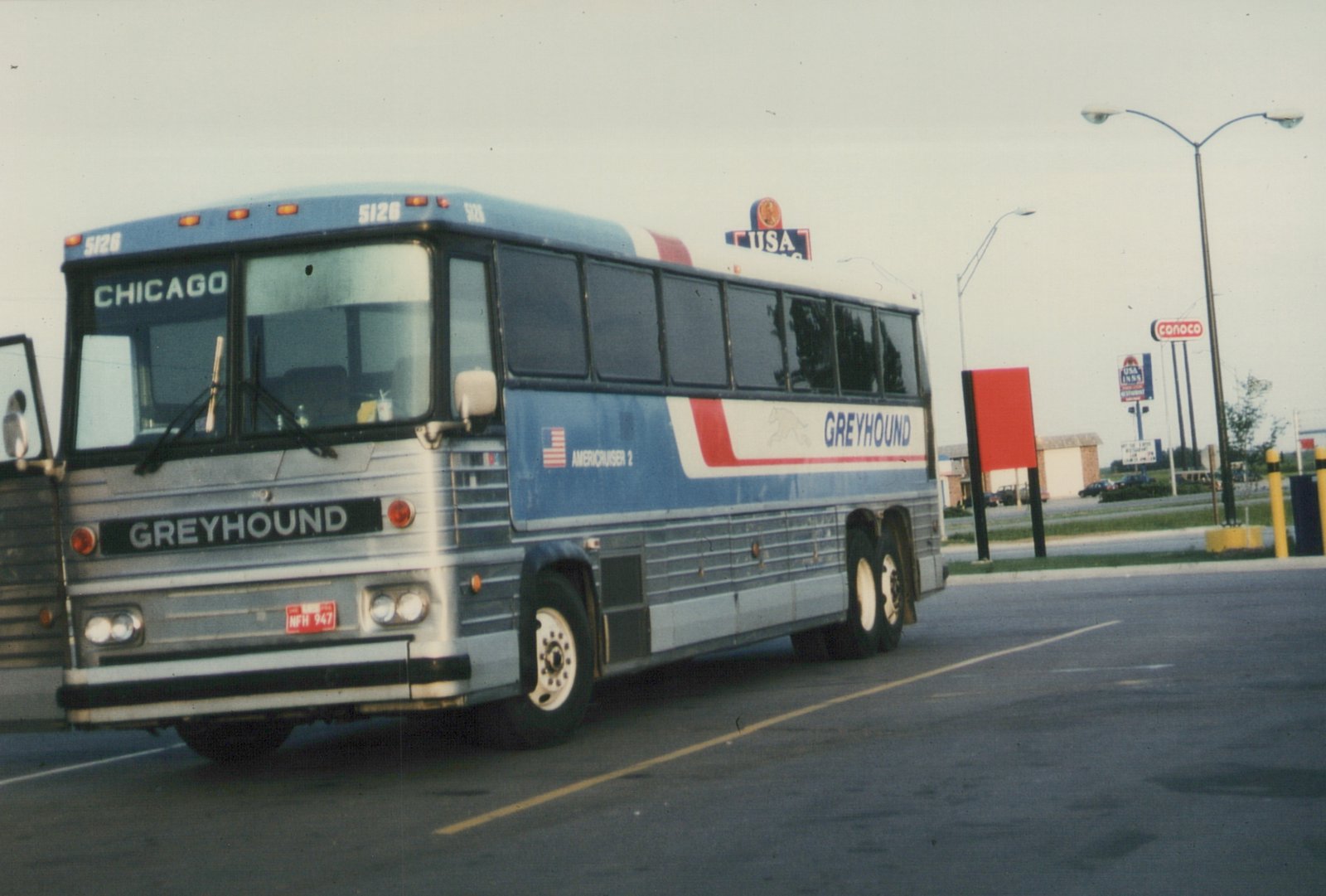 A front three-quarter view of a Greyhound bus, number 5126, with 'CHICAGO' on the destination window, parked in a lot with USA INNS and CONOCO signs in the background.