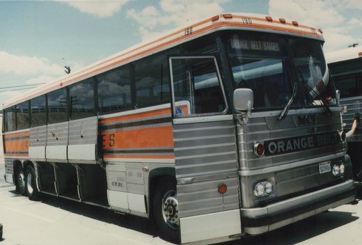 A silver and orange MCI MC9 bus with the fleet number 190 and the name Orange Belt Stages on the front, parked on a paved lot with a clear blue sky above.