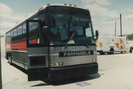 A silver, black and red Panhandle Trailways bus with a destination sign reading 'LOS ANGELES' is parked on a paved lot, with a white delivery truck advertising Corona Extra visible in the background.