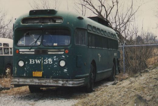 A three-quarter front view of a dark green, weathered bus with the number BW 35 on its front and side, parked on a snowy, muddy lot next to a chain-link fence and bare trees.