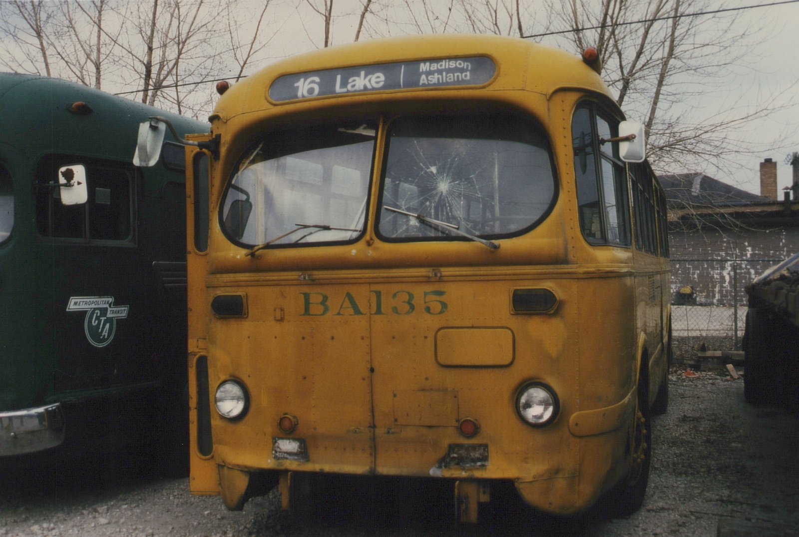 Front view of a yellow CTA bus, number BA135, with a broken windshield and a '16 Lake | Madison Ashland' destination sign, parked next to a green CTA bus in a gravel yard with bare trees.