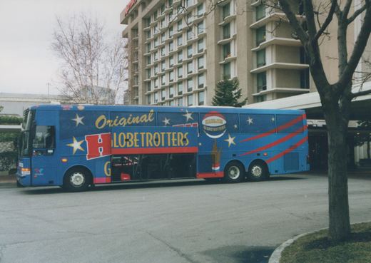 Side view of a blue motor coach wrapped in a Harlem Globetrotters design, parked on a paved area with a large building and bare trees in the background.