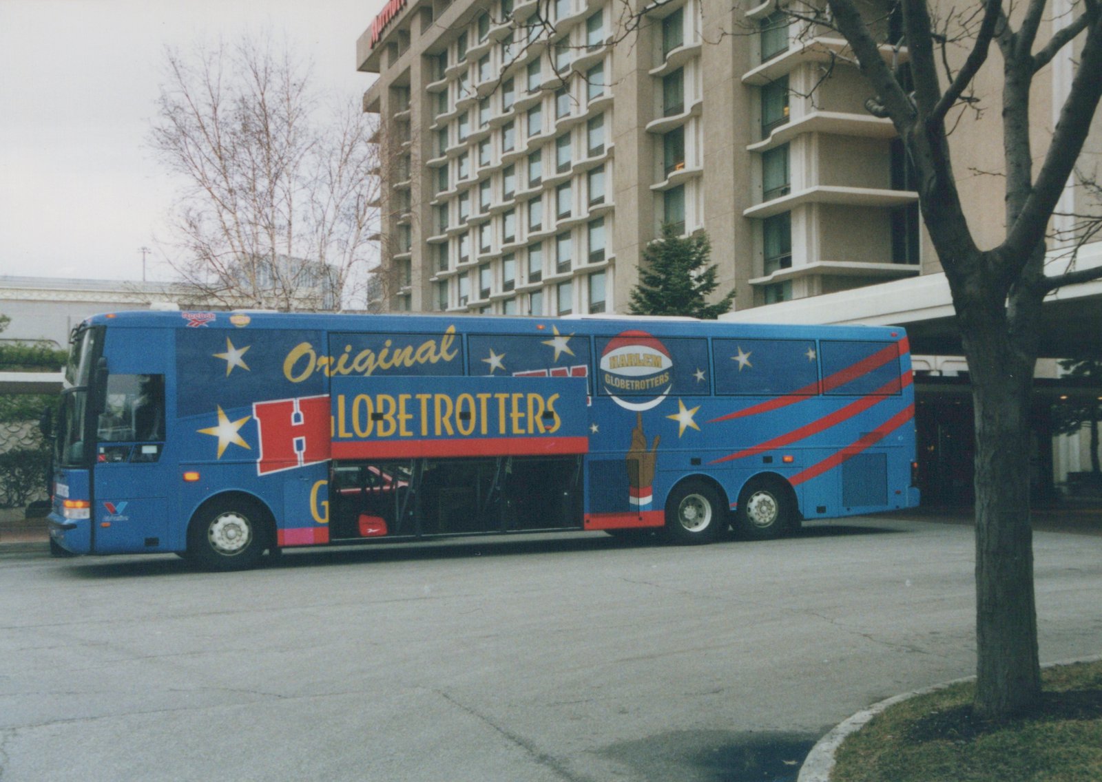 Side view of a blue motor coach wrapped in a Harlem Globetrotters design, parked on a paved area with a large building and bare trees in the background.