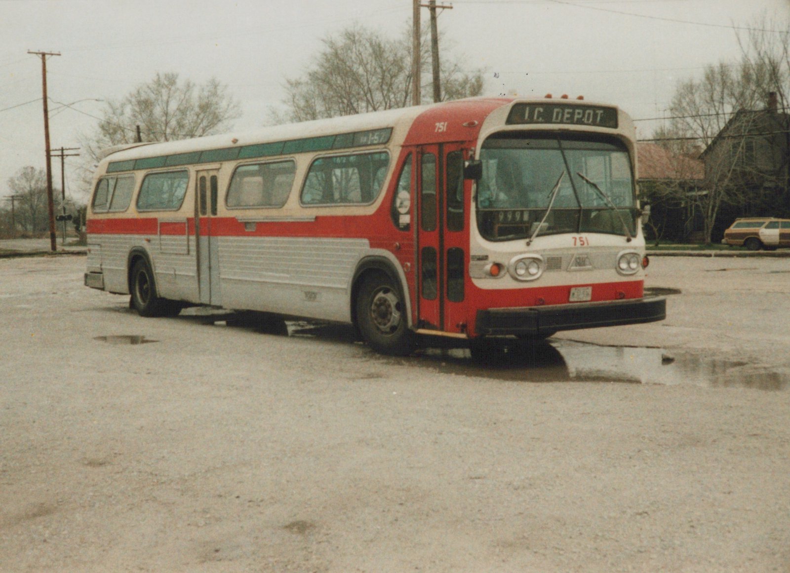 A red, white and green Illinois Central motor bus numbered 751 is parked on a wet, paved lot on an overcast winter day, with a station wagon and bare trees in the background.