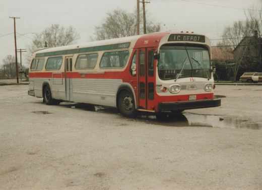 A red, white and green Illinois Central motor bus numbered 751 is parked on a wet, paved lot on an overcast winter day, with a station wagon and bare trees in the background.