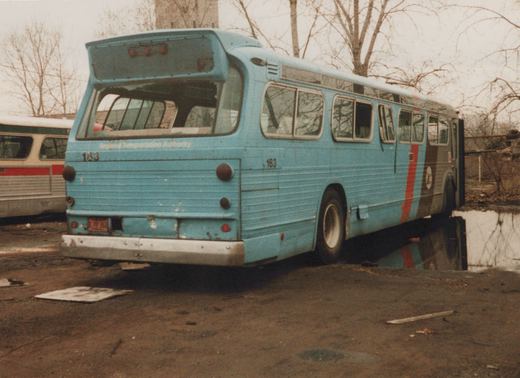 Rear three-quarter view of a light blue Regional Transportation Authority bus, number 163, parked in a muddy yard with bare trees in the background.
