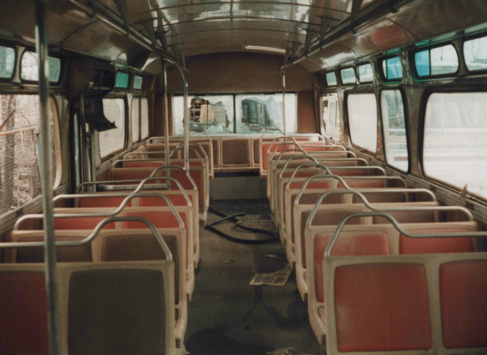 The interior of an empty, damaged bus with red seats, litter on the floor, and a broken windshield, viewed from the rear looking forward.