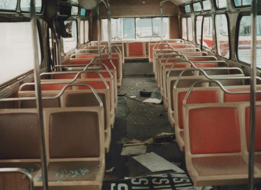 The interior of a damaged bus with shattered windows, littered floor, and rows of red and brown seats, viewed from the rear looking forward.