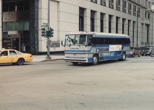 A white and blue Keeshin bus, number 824, is driving on a city street with a yellow taxi and other cars nearby, at the corner of Franklin Street and Wacker Drive in Chicago.