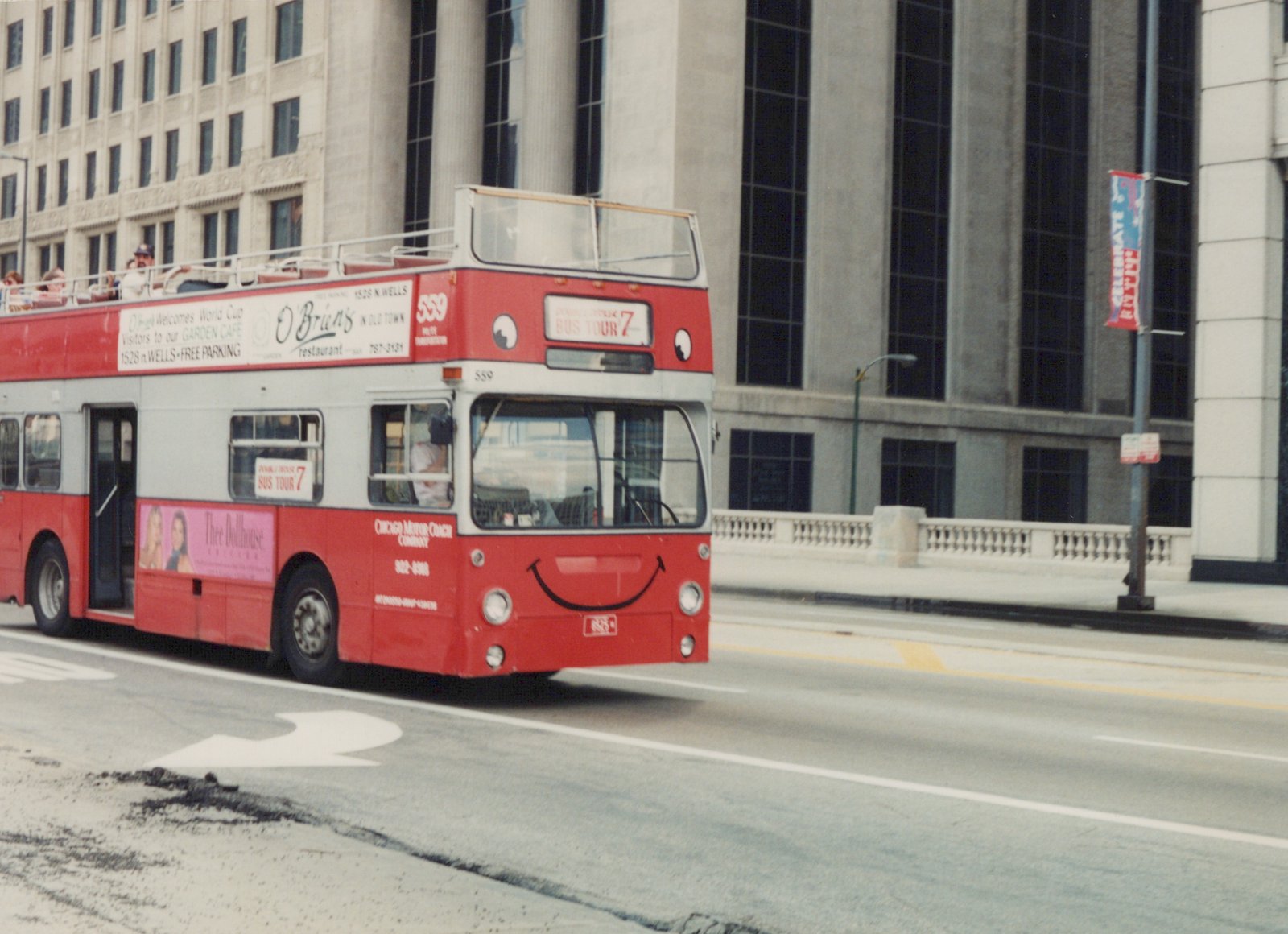 A red and white double-decker tour bus with the number 559 is driving on a city street. Passengers are visible on the open top deck. The bus has a black smiley face on its front and 'CHICAGO MOTOR COACH COMPANY' written on its side. A banner in the background reads 'CELEBRATE '94'.