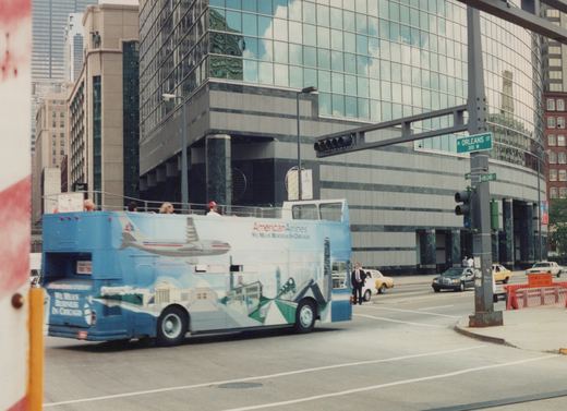 A blue and white double-decker bus with 'American Airlines' lettering is turning a corner on a city street, with passengers on the open top deck and a modern glass building behind it.