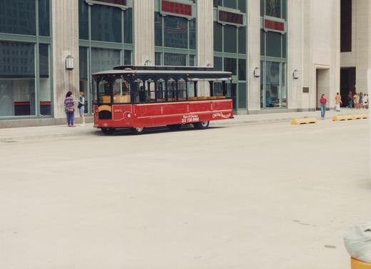 A red trolleybus labeled 'Chicago Trolley' and number 990 is parked on a city street with people nearby and a large building in the background.