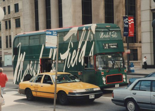 A green double-decker CTA bus with a Marshall Field's ad is on a city street, stopped near a bus stop sign, with a yellow taxi and a silver sedan in the foreground and a 'Chicago '96' banner on a pole in the background.