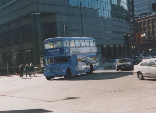 A blue double-decker bus with airline ads drives down a city street, with pedestrians and other cars nearby.