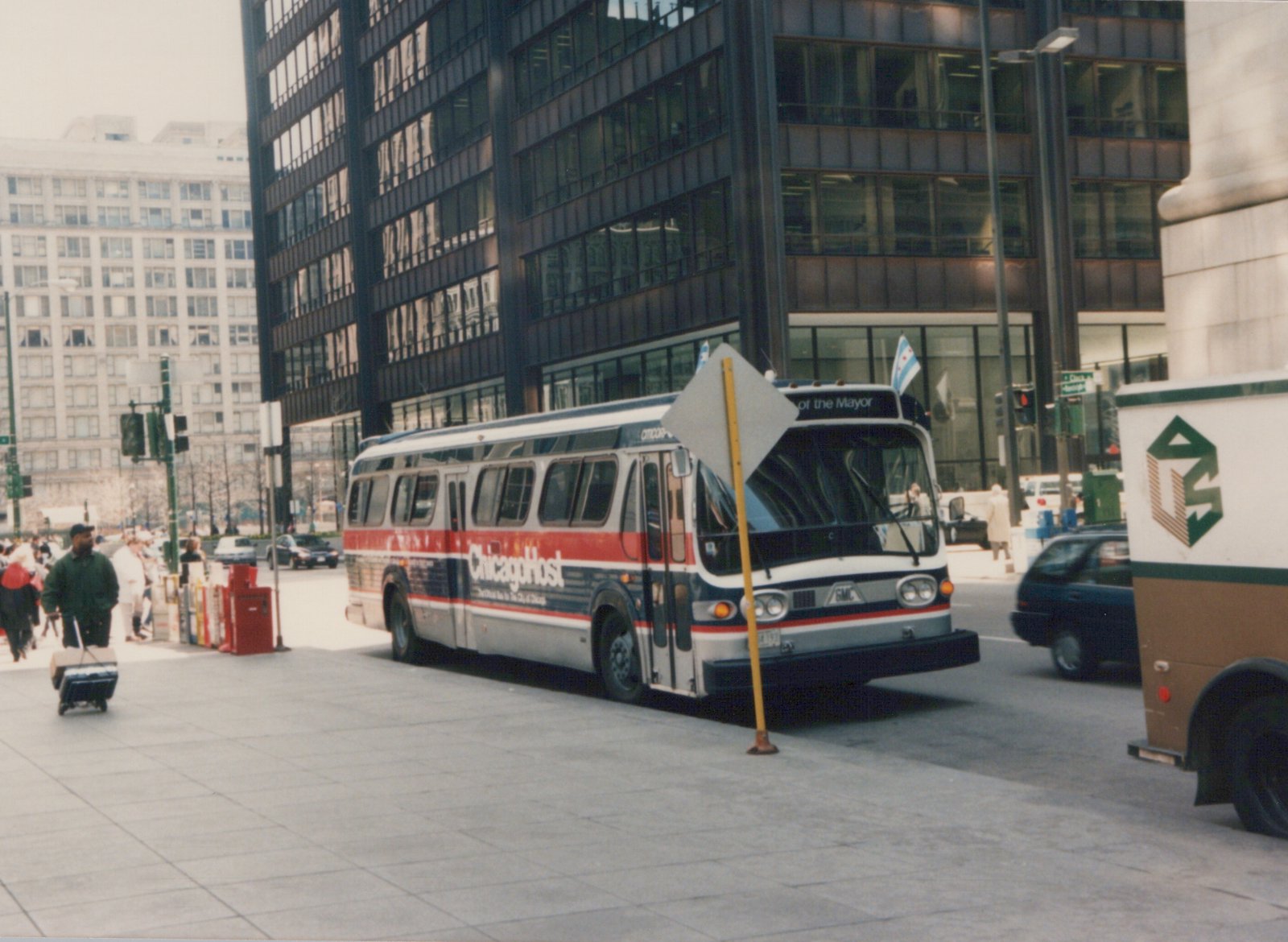 A CTA bus in red, white and blue livery with 'Chicago Host' lettering is parked on a city street with tall buildings and pedestrians nearby.