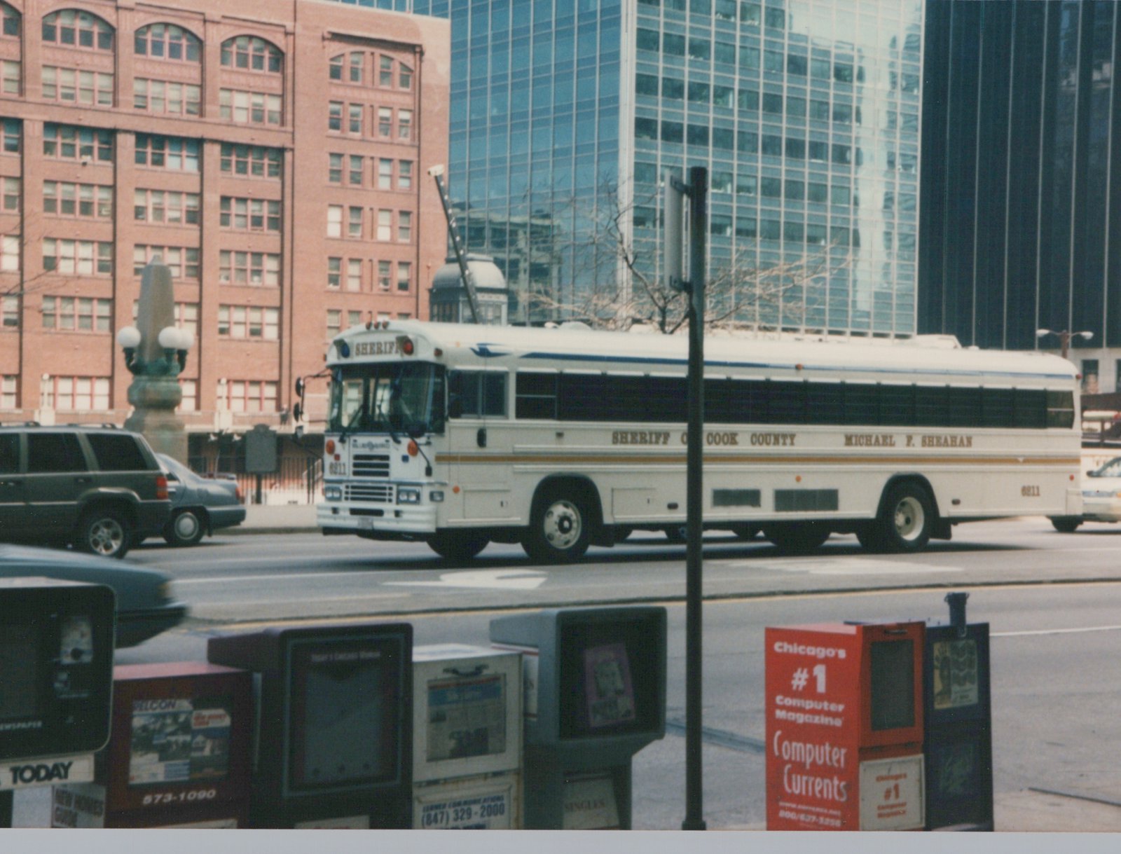 A white Cook County Sheriff's bus, number 6811, is parked on a city street with tall buildings in the background and newspaper boxes in the foreground.