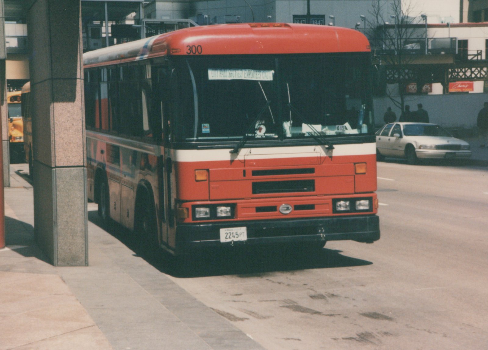 Front three-quarter view of a red and white CTA bus number 300 on a city street, with a destination sign reading 'CITY HALL SHUTTLE / EMPLOYEES ONLY', and a white car in the background.