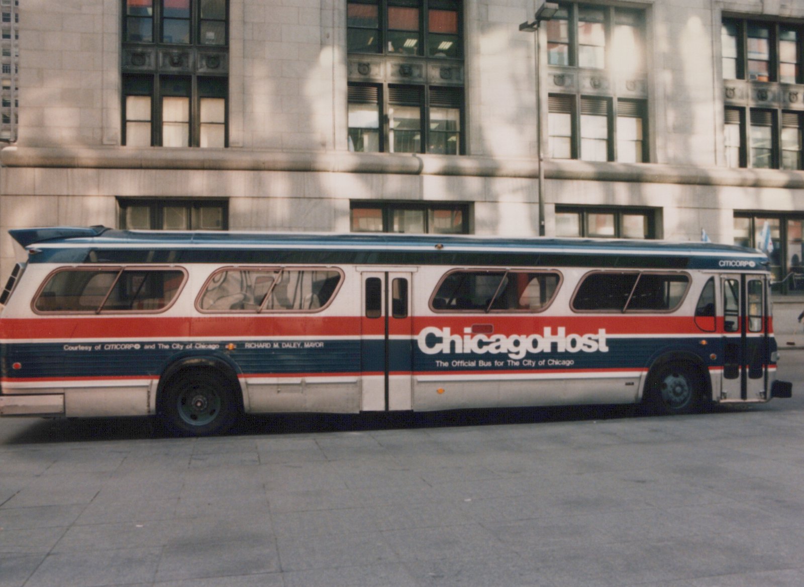 A white, red, and blue bus with 'ChicagoHost' written on the side, parked on a paved area in front of a large stone building.