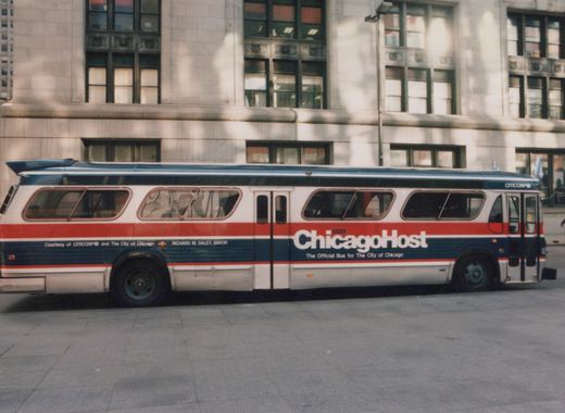 A white, red, and blue bus with 'ChicagoHost' written on the side, parked on a paved area in front of a large stone building.