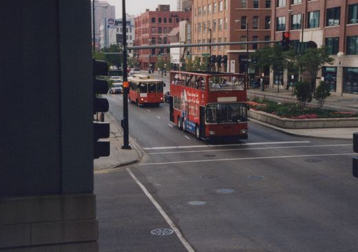 A red double-decker bus with 'Chicago Motor Coach Company' lettering and the number 568 is turning at an intersection with a single-decker bus behind it, in a city street scene with buildings and pedestrians.