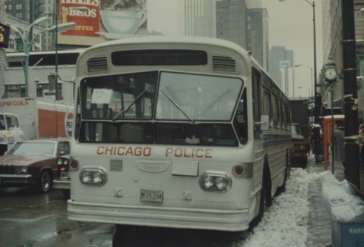 Front view of a white Chicago Police bus, number 007, parked on a snowy Chicago street with buildings and a Pepsi truck in the background.