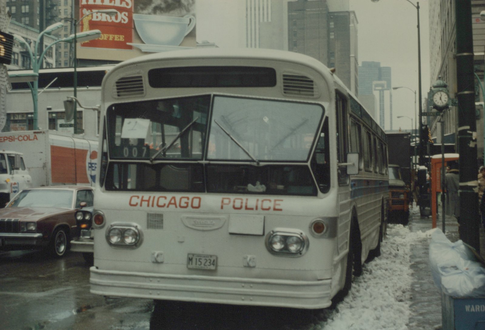 Front view of a white Chicago Police bus, number 007, parked on a snowy Chicago street with buildings and a Pepsi truck in the background.