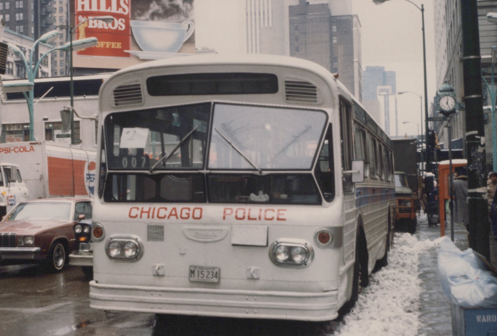 A white Chicago Police bus with the number 007 on its front sign is parked on a snowy city street, with buildings and a Pepsi delivery truck visible in the background.