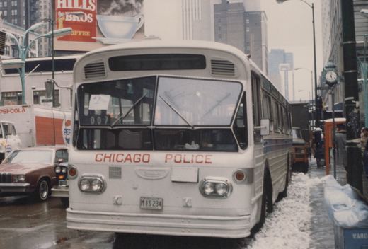 A white Chicago Police bus with the number 007 on its front sign is parked on a snowy city street, with buildings and a Pepsi delivery truck visible in the background.