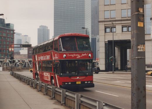 A red double-decker bus with 'Chicago Motor Coach Company' and 'Luxury Service with a Smile!' lettering is driving on a city street with skyscrapers in the background.