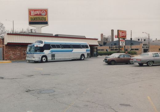 A white and blue Pace bus is parked in a cracked asphalt lot next to a Wendy's restaurant and a bowling alley, with two 1980s cars nearby.