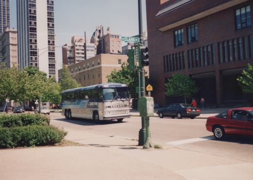 A blue, white, and silver Keeshin motor bus is turning a corner on a city street, with a street sign reading 'W CLARK ST' in the background and other cars nearby.