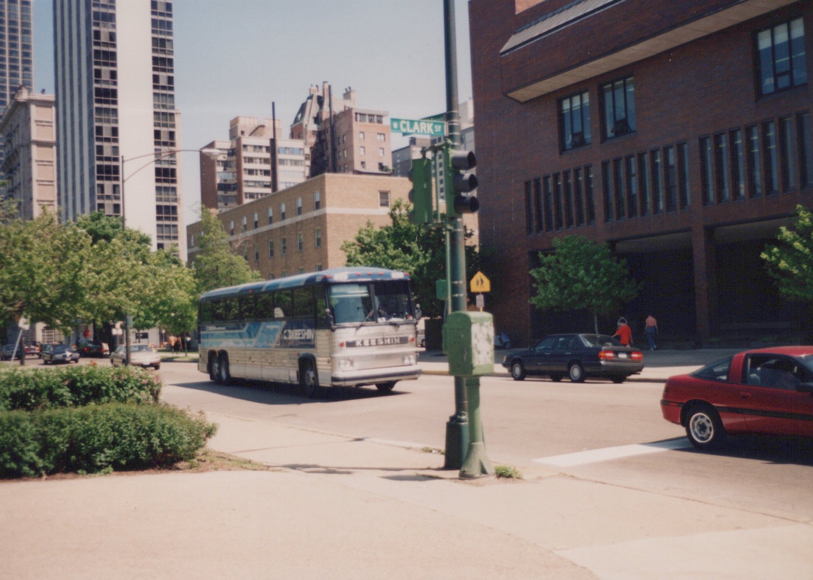 A blue, white, and silver Keeshin motor bus is turning a corner on a city street, with a street sign reading 'W CLARK ST' in the background and other cars nearby.