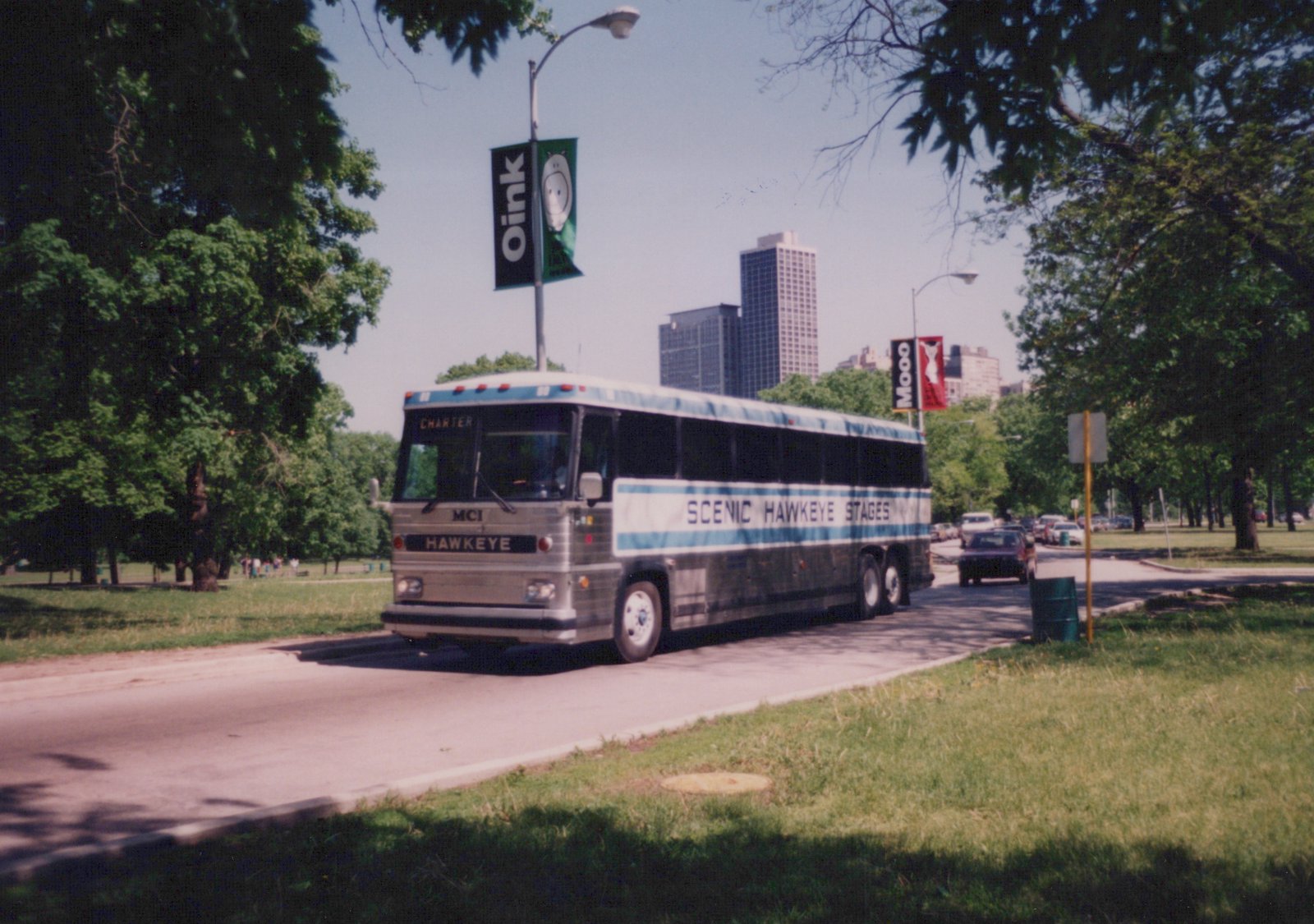A silver and blue motor coach labeled 'Scenic Hawkeye Stages' travels down a road lined with trees and urban banners, with tall buildings in the distance.