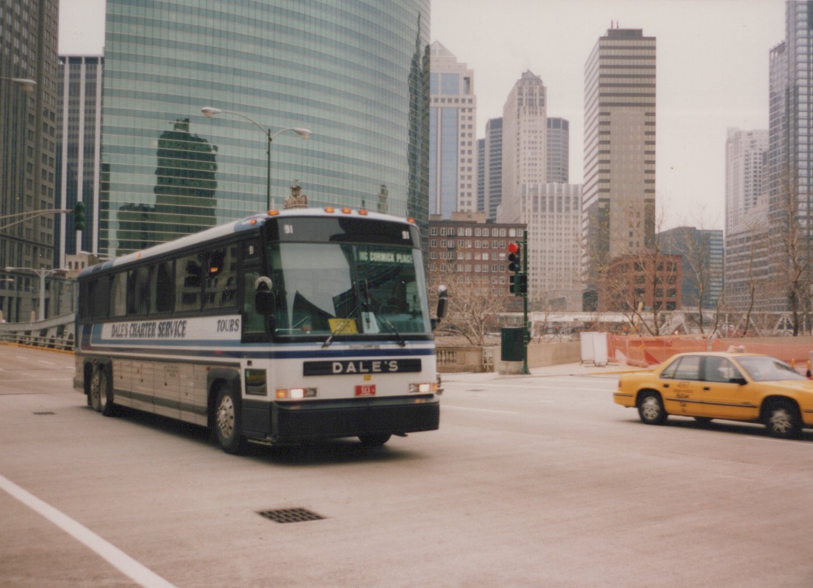 A white and blue charter bus with the number 91 on the front, labeled 'Dale's Charter Service,' is driving on a city street with tall glass buildings behind it and a yellow taxi nearby.